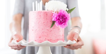 A beautiful pink cake topped with pink and white flowers and lit candles is held on a white cake stand.