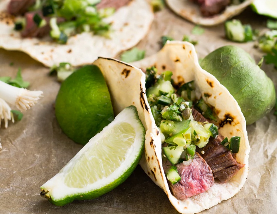 A soft shell taco containing steak and salsa verde is propped up by limes.
