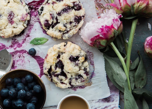A few blueberry scones sit on a cooking surface between a small bowl of blueberries and 2 white flowers.